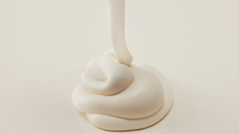 A high-angle close-up shot of a rich, whipped luxury face cream in a heavy glass jar, resting on an ivory silk cloth with soft, directional morning light highlighting its creamy texture