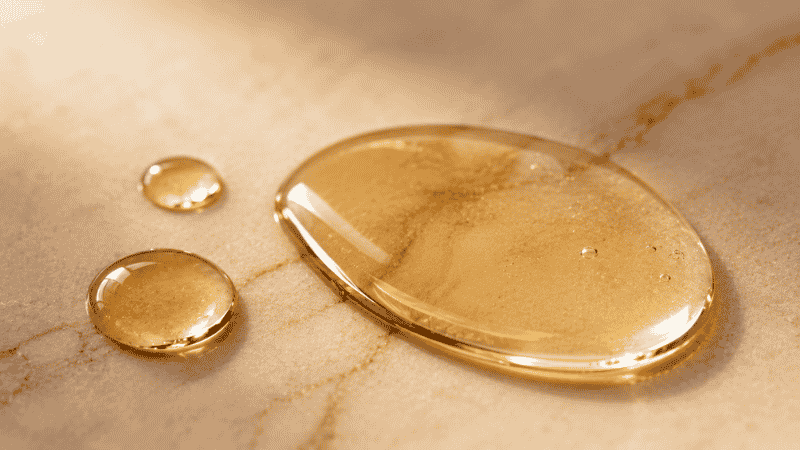 Close-up macro of golden squalane oil drops on marble — the lightweight skin-identical texture that gives squalane its skin benefits