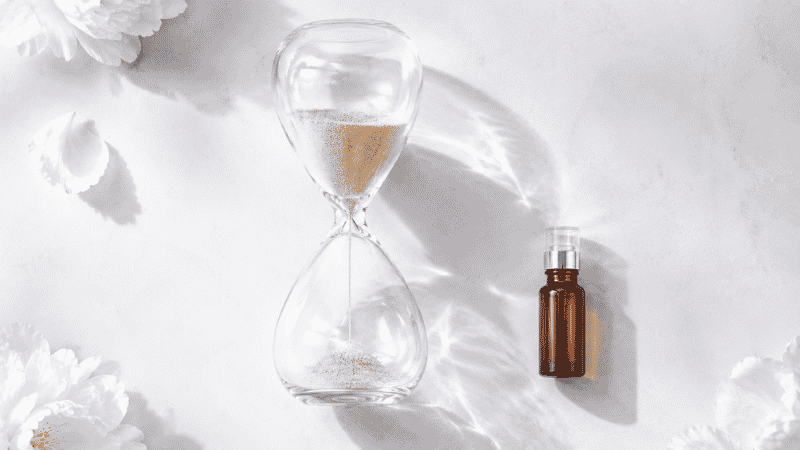 Glass hourglass with gold sand beside luxury skincare vial on white — skin changes in your 30s including collagen decline of 1% per year and the case for early intervention